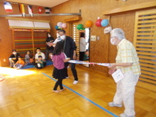 広いスペースで、青いテープが床に貼られ、中央で子どもが棒を持ち、両端を大人が支えている様子が写された写真