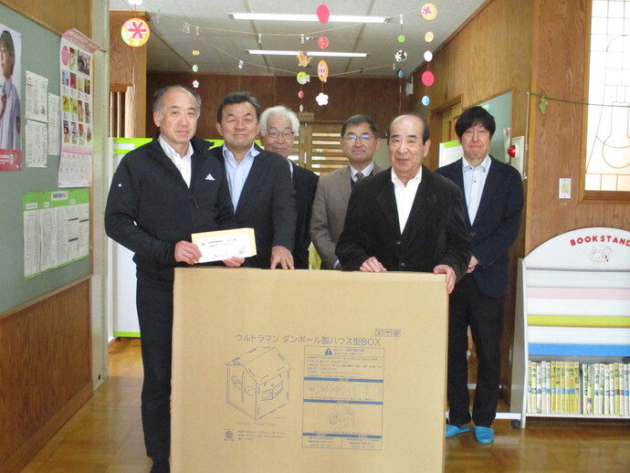 複数のスーツ姿の人物が並び、前方には「ウルトラマン ダンボールおうちBOX」と記載された大型段ボール箱が置かれ、左側の人物が封筒を持っている写真