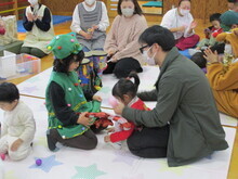 星柄のシートの上で、緑のクリスマスツリー風衣装を着た子どもが座り、赤い服の子どもと隣の人物が向かい合って遊んでいる室内の写真