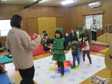 広い木目調の室内で、ツリーのような緑色の衣装を着た女の子や幼い子どもたちが星柄のマットの上で周囲の大人たちに見守られながら交流をしているクリスマス会の様子の写真