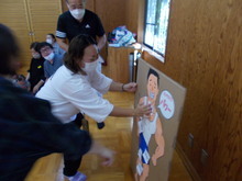 複数の人が集まり、中央では白い服を着た人物が段ボールに描かれた運動選手のイラストにパーツを貼り付ける保護者競技をしている様子が写された写真