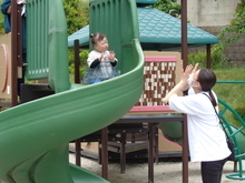 緑色の曲線状の滑り台の上に子どもが座り、下から白い服を着た人物が両手を上げて向かい合って遊んでいる写真