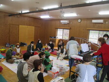 星柄のシートの周りに複数の人々が座り、中央では子どもたちが遊んでおり、右側では赤い服の人物が電子ピアノを演奏している写真