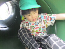 緑色の帽子をかぶった子どもが、緑色の筒状の滑り台の中で片手を広げて座っている様子が写っている写真