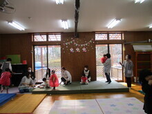 広い室内で、数名の保護者や保育士が見守る中幼い子どもたちが赤い色の衣装を着て遊戯をしている様子の写真