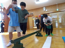 中央に緑色の服を着た子どもが平均台を渡り、横で大人が支えており、後方でも別の子どもが保護者に手を添えられながら平均台に挑戦をしている様子が写された写真