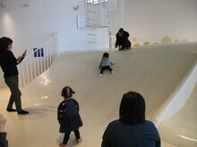 白い室内遊具の滑り台で複数の子どもとが大人が遊んでいる明るい空間の写真