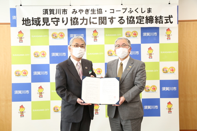 「須賀川市 みやぎ生協 コープふくしま 地域見守り協力に関する協定締結式」と書かれた横断幕を背景にスーツを着たマスク姿の関係者の男性2人が締結書を一緒に持ち記念撮影を行っている写真
