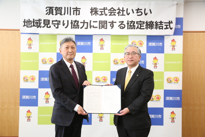 「須賀川市 株式会社いちい 地域見守り協力に関する協定締結式」と書かれた横断幕を背景にスーツ姿の関係者の男性2人が締結書を一緒に持ち記念撮影を行っている写真