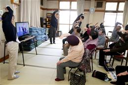 和室で、椅子に座ったたくさんの高齢者の参加者が、テレビモニターに映る映像と前に並んだ指導員を手本にしながら、一斉に腕を高く伸ばして上体を横に倒すストレッチに取り組んでいる様子の写真