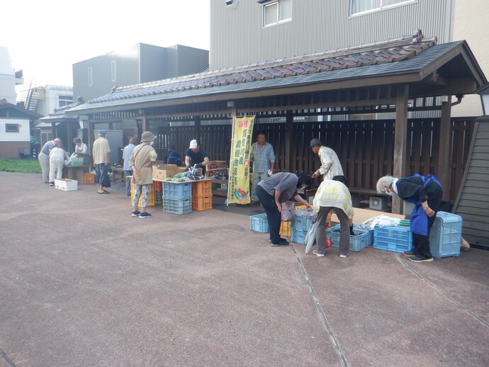 屋外の屋根付きのスペース前に黄色いのぼりが立てられ、野菜などが入ったたくさんの水色やオレンジ色のコンテナが並ぶ青空市場が来場者や生産者で賑わっている様子の写真