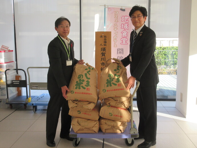 紫色の台車に8つの米袋が積まれ、両側に立ったスーツ姿の男性2人が、それぞれ米袋の上を持ち上げている、出品米の寄付の様子の写真