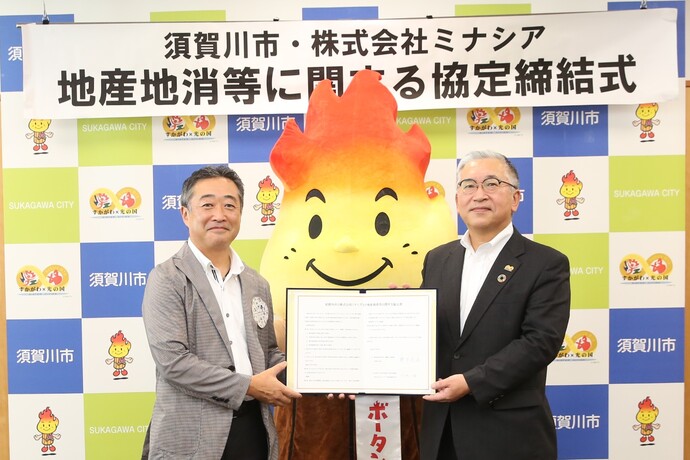 「須賀川市・株式会社ミナシア地産地消等に関する協定締結式」の横断幕が設置された須賀川市のバックパネルの前で、赤い炎のキャラクター「ボータン」のマスコットを挟んで立った男性2人が左右から協定書を持って写した記念写真