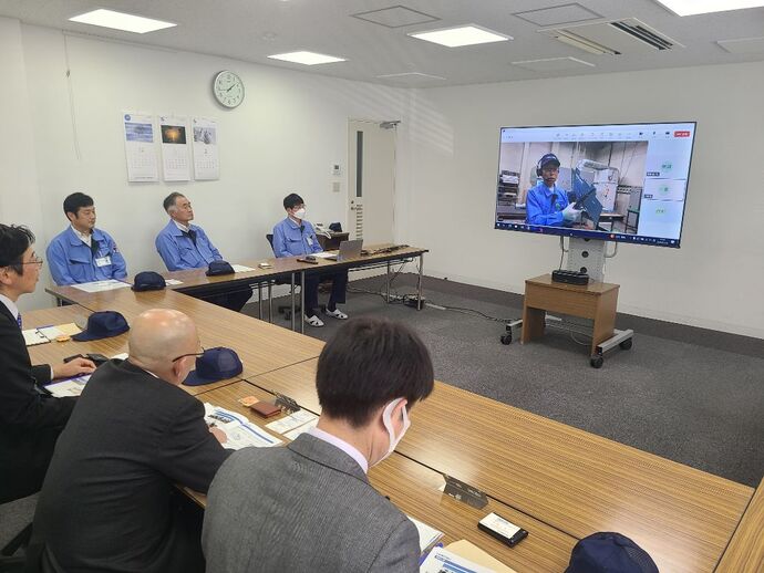 会議室の長机に座ったスーツや作業着姿の男性たちが、大型モニターに映し出された青い作業着で部品を手にした人物によるリモート説明を真剣な表情で見つめている事業説明の写真