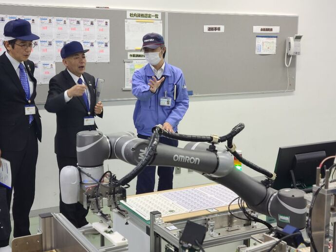 青い作業着を着た担当者が「OMRON」と書かれた産業用ロボットアームの動作について、青い帽子とスーツを着用した二人の視察者に身振り手振りを交えて説明している写真