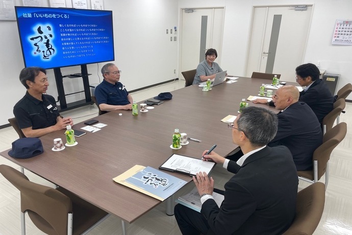 茶色の長机を囲んでスーツ姿の参加者が着席し、壁の大型ディスプレイに青い背景の杜温泉はなきつくりのスライドが表示され意見交換を行っている様子の写真