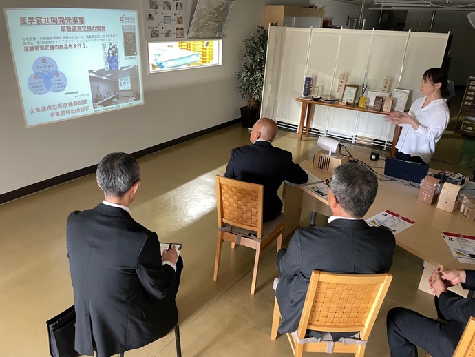 「産学官共同開発事業」と書かれたスライドを前に、白いシャツを着た樽川千香子 代表取締役社長が説明を行い、スーツ姿の男性たちが熱心に耳を傾けているプレゼンテーション中の写真