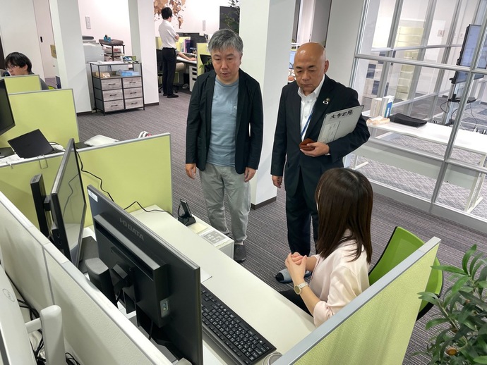 明るいオフィス内で、黄緑色のパーティションで区切られたデスクに座る女性社員に、スーツ姿の男性2人が立って話しかけている様子の写真