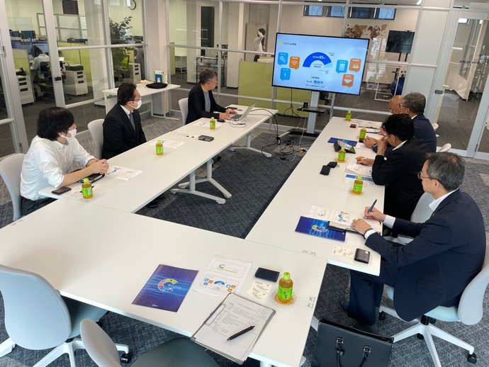 白いコ字型テーブルを囲んでスーツ姿の参加者がマスクを着用して着席し、壁のモニターにプレゼンテーション資料が投影されている会議室の写真