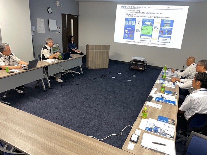 会議室で複数の参加者がコの字型に設置された長机に着席し、ノートパソコンや資料を前にプロジェクターで表示されたIT機器の管理ソリューションに関するスライドを見ながら説明を受けている様子の写真