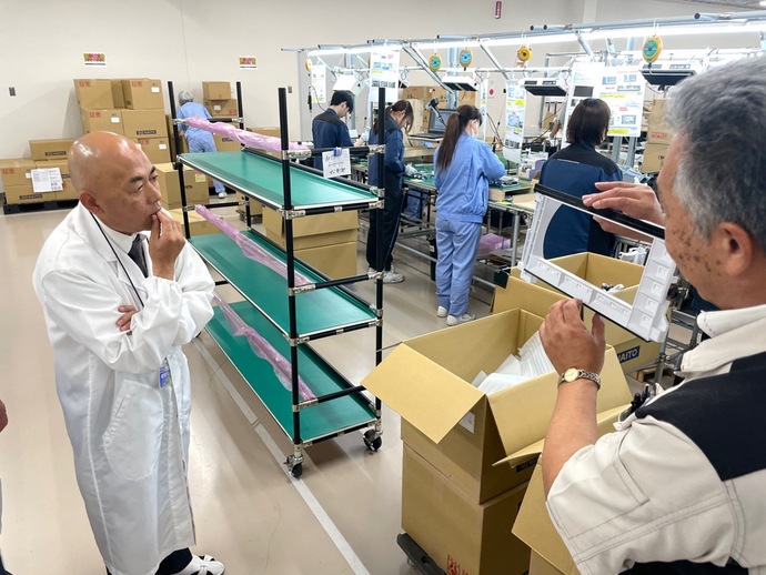 工場の組立ラインで複数の作業者が青や濃紺の作業着を着て製品を組み立てており、手前には緑色の棚と段ボール箱が置かれ、佐藤事業部長が液晶ユニット製造を男性に解説している様子の写真