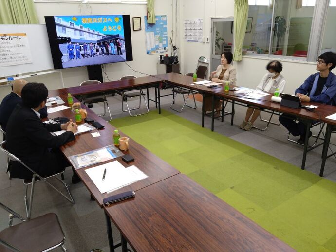 緑色のカーペットが敷かれた会議室で、コの字型に配置された長机を囲んで座る数名の男女が、壁際に設置された大型モニターに映し出された資料を見ている写真