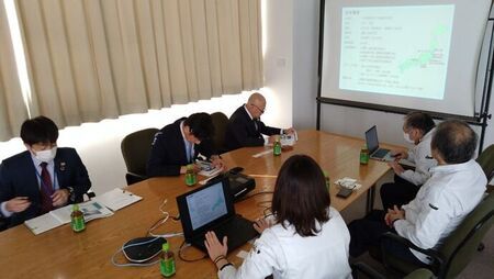 会議室にて、日本地図や箇条書きのテキストが映し出されたスクリーンの前で、長テーブルに座る男女6名の参加者が手元の資料やノートパソコンを確認しながら会議を行っている様子を捉えた写真