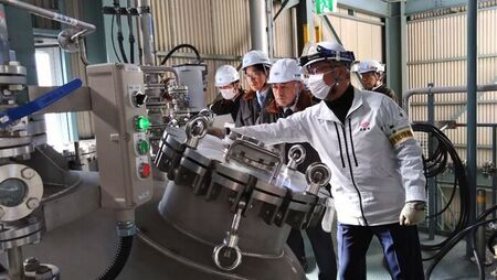ヘルメットとマスクを着用した社員の男性が、工場内の複雑な配管が繋がる大きな金属製タンクの蓋部分を指差しながら、ヘルメットをかぶった見学者の男性たちへ説明をしている様子の写真