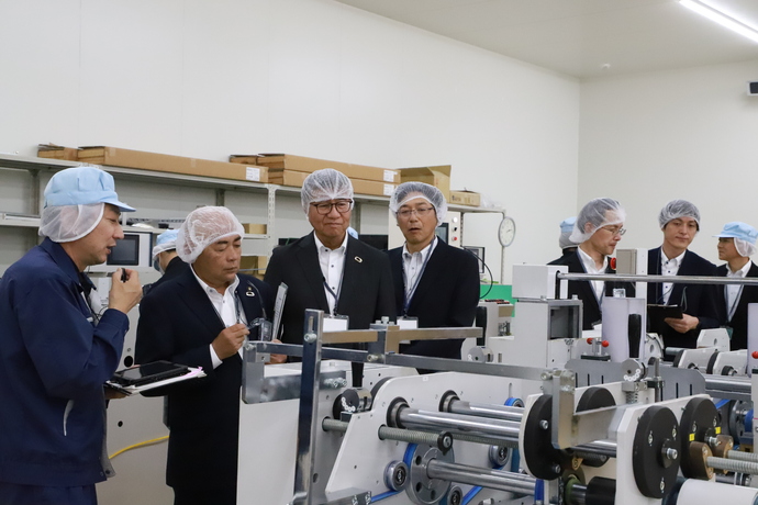 白い衛生キャップを着用したスーツ姿の視察者たちが、工場内の製造ラインの機械設備を視察し、作業着姿の担当者から説明を受けている様子の写真