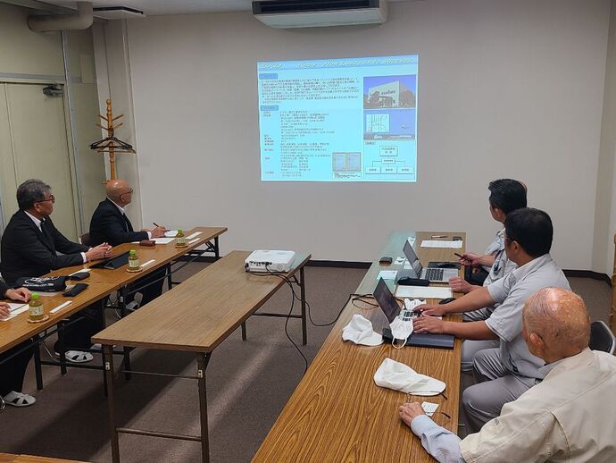 木製長机が並ぶ会議室で、スーツ姿の男性と作業服姿の参加者がプロジェクターに投影された資料を見ながら会議を行う様子の写真