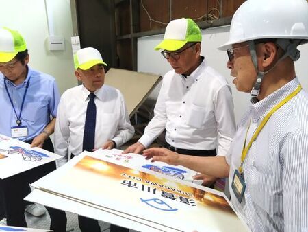 白いヘルメットを被った男性が「須賀川市」と記載されたパネルを指さしながら、黄色いキャップを被った3人の男性たちに説明を行っている視察風景の写真