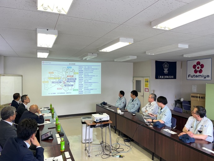 会議室で複数の人が向かい合って置かれた長机の席に座り、前方の壁に地図と地域別情報が表示された資料を投影しながら説明を受けている様子の写真