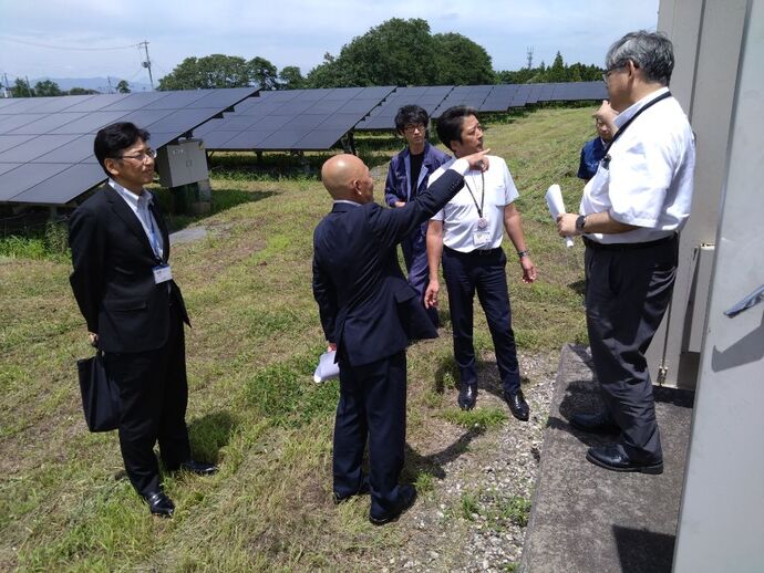 広大な敷地に整然と並ぶ多数のソーラーパネルを背景に、スーツや作業服を着用した数名の男性たちが集まり話をしている様子の写真