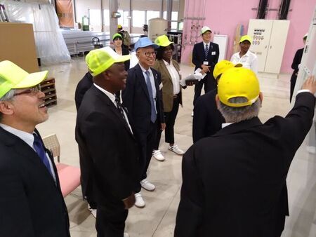 ピンク色の壁がある工場内で、黄色いキャップを被ったザンビア駐日大使やスーツ姿の男性たちが、案内役の話に笑顔で耳を傾けながら施設内を見学している様子の写真
