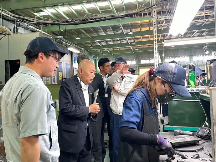 工場内で手袋を着けた人物が金属部品の加工を行っている様子を後方からスーツを着た男性たちが見学し、説明を受けている様子の写真