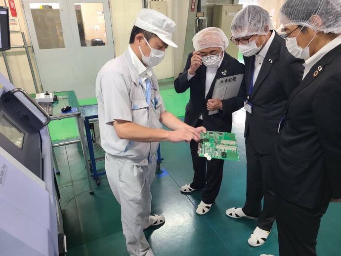 緑色の床の工場内で、白い作業着と帽子の従業員が電子基板を持ち、マスクと衛生キャップを着用したスーツ姿の視察者たちに製品を説明する様子の写真