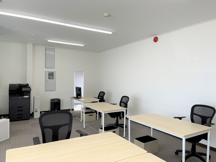 白を基調とした清潔感のある室内に、木目調のデスクと黒いメッシュチェアが整然と並び、左奥には大型の複合機が配置された、コワーキングスペースCOCO-LABOの室内写真