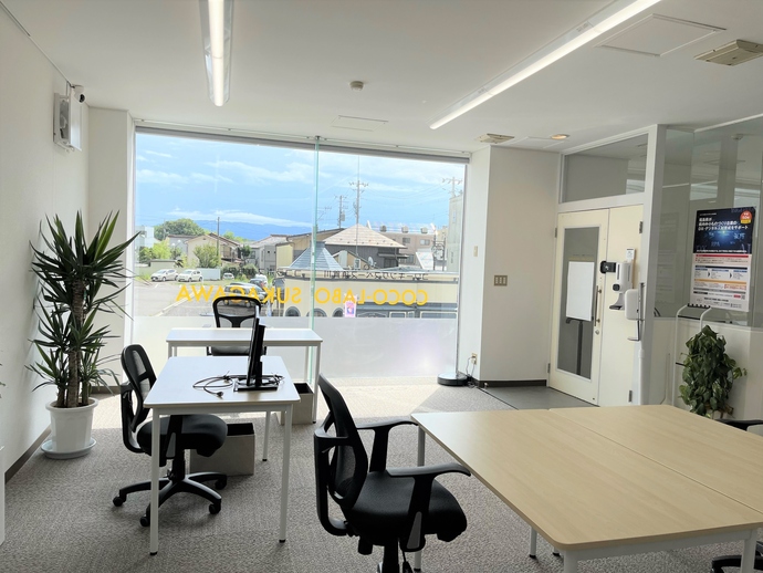 大きな窓から明るい光が差し込む室内に、白いデスクや黒いワークチェアが配置され、観葉植物の緑がアクセントを添える、清潔感あふれるモダンで開放的なコワーキングスペースCOCO-LABOの室内写真