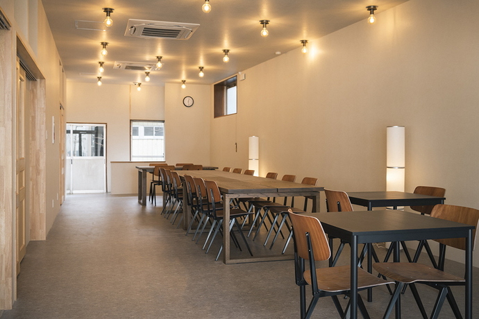 温かみのある電球色の照明が天井に並び、木目調の長テーブルや黒い小型テーブルにモダンな椅子が整然と配置された、シンプルで落ち着いた雰囲気の共有スペースの写真