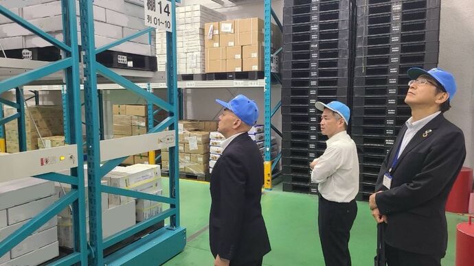 青い帽子を被った3人の男性が、倉庫内で高く積み上げられた段ボール箱やプラスチックパレットが並ぶ大型の移動式棚を、真剣な表情で見上げながら視察を行っている様子を捉えた写真