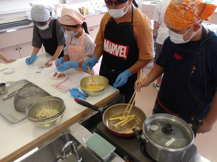 調理室でエプロンと三角巾を着けた4人の児童が、手袋をして、材料や道具が並んだ作業台で下準備をしたり、その隣に置かれたフライパンでパンの耳を焼いたりと、調理を行っている様子の写真