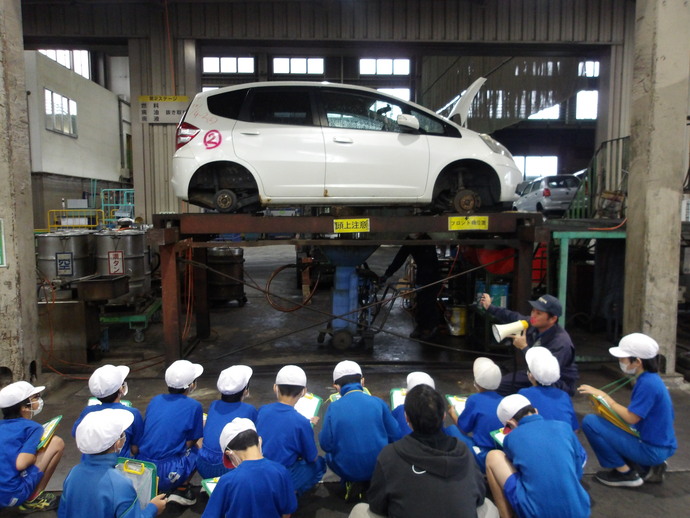 自動車整備工場の屋内で、リフトに載せられた白い車両がタイヤを外した状態で整備されており、前方では青い服と白い帽子を着た子どもたちが床に座ってノートを取りながら見学し、説明役の人物がメガホンを使って話している様子を写した写真