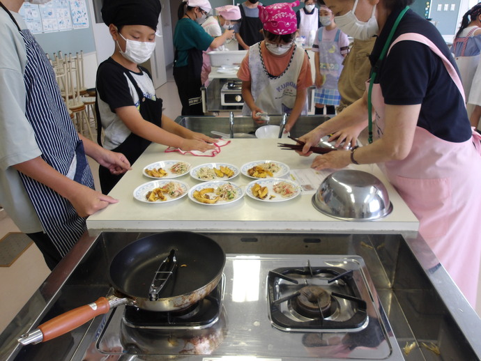調理室でエプロンと三角巾を着けた児童たちと講師の女性がグループに分かれ作業をしており、手前のグループの調理台では、中央の白いカウンターに料理が盛り付けられた皿が6枚並び、3人の児童と講師の女性が配膳や調理の片づけを行っている様子の写真