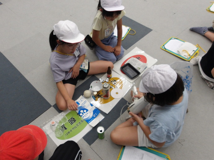 床に座った児童が白い帽子をかぶり、空き缶や瓶、トレーなどのごみを分別する学習を行い、ノートや資料を広げている様子の写真