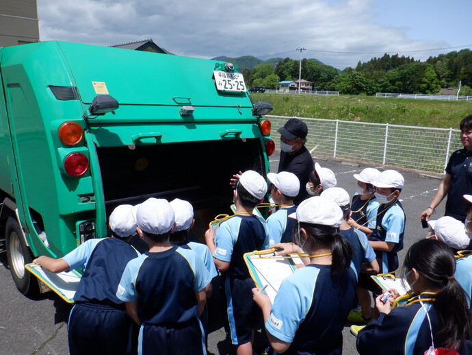 屋外で緑色のごみ収集車の後部を囲むように、白い帽子と青い運動服を着た子どもたちが並び、手には黄色いバインダーを持って説明を聞いている様子が写された写真