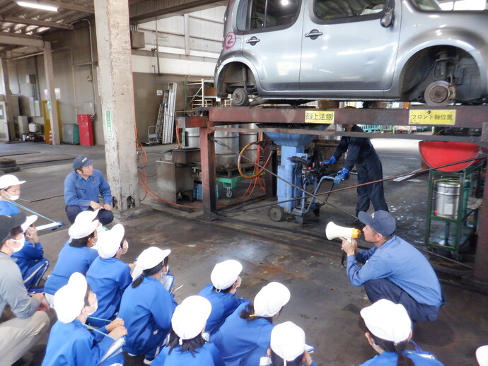 自動車整備工場のような屋内で、リフトに載せられたシルバーの車両の下で作業員が工具を使って整備を行っており、前方では青い作業服と白い帽子を着た子どもたちが床に座って見学し、説明役の人物がメガホンを持って話している様子を写した写真