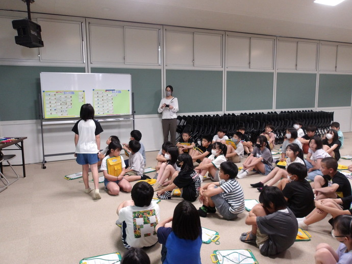 教室内で多数の児童が床に座り、前方のホワイトボードに貼られた紙を見ながら学習活動を行っており、中央には一人の児童が立って発表し、後方には大人が立って様子を見守っている様子の写真
