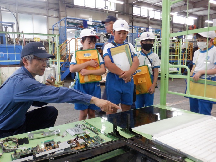 作業服を着た男性が手元の電子基板やパネルを指差しながら説明し、白い帽子と青いズボン姿の小学生4人が真剣な表情でその様子を見学している写真