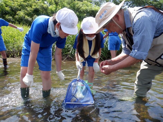 すかがわの川の中に立ち、青い網を手にした白い帽子と青いジャージ姿の2人の子供たちが、男性の手元を興味深そうに覗き込みながら、観察を行っている様子の写真