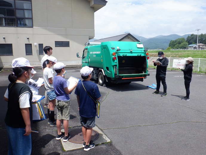 ゴミ収集車を前に、白い帽子を被り探検バッグを手にした小学生たちが、説明を行う作業員の話を真剣な表情で聞きながらメモを取っているESD環境教育の様子の写真